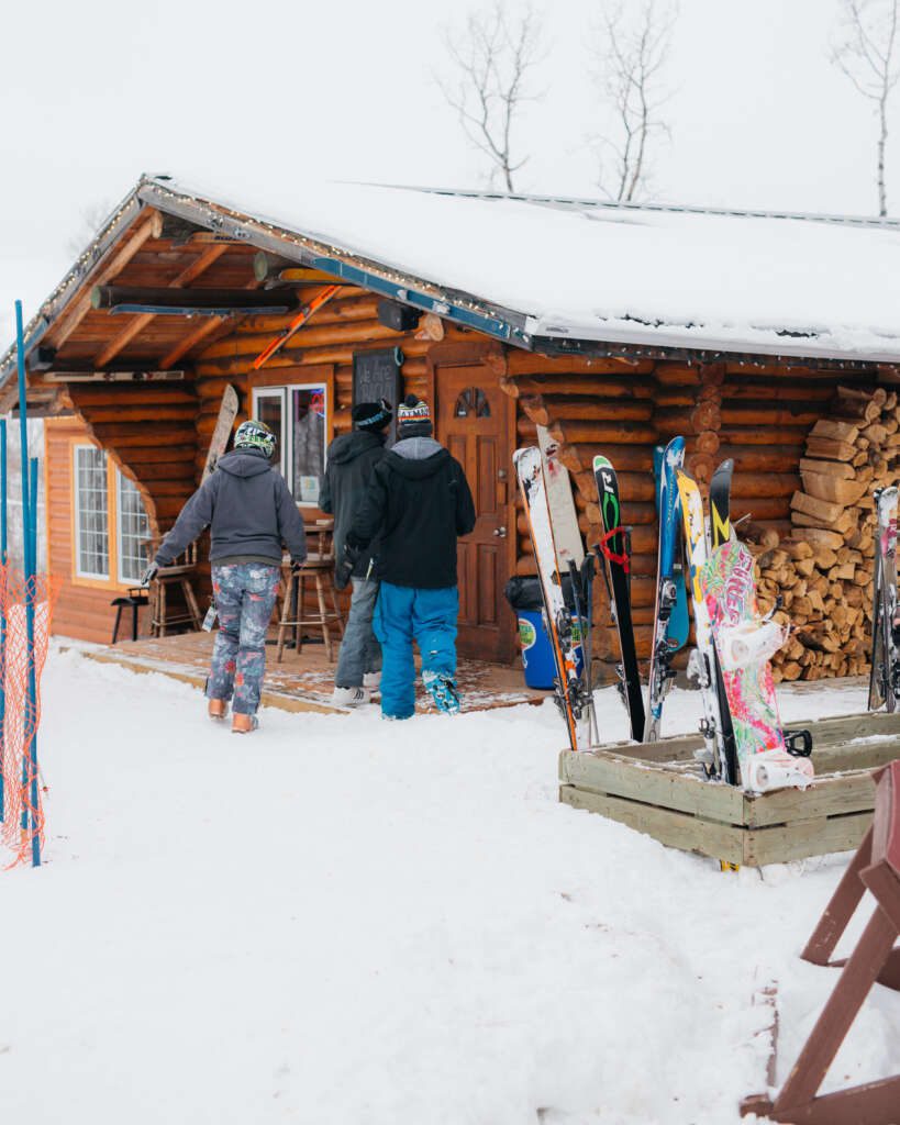 three people walking into a log cabin with skis and snowboards to the side