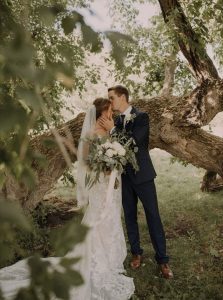 groom kissing bride on the forehead in front of a tree