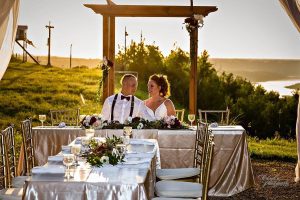 Newlyweds sitting a table at their reception with the altar and a sunset behind them